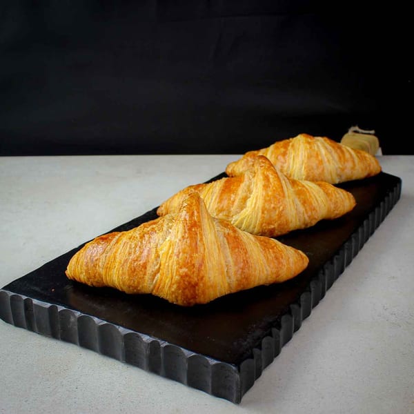 Three golden-brown croissants from iCafe de Paris, arranged on a dark, textured serving board.