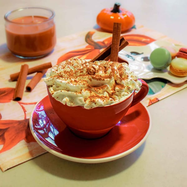Pumpkin spice latte with whipped cream and cinnamon sticks, with macarons and candle in the background.