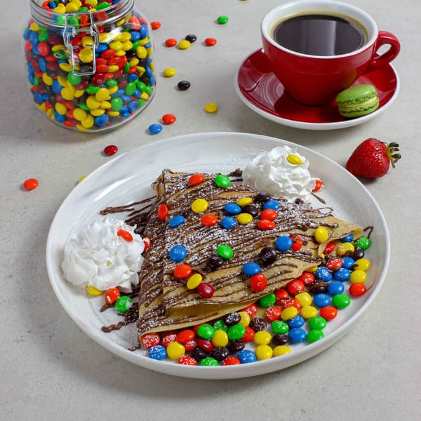 Chocolate crepe with M&M's, whipped cream, powdered sugar, a red coffee cup, and a green macaron.