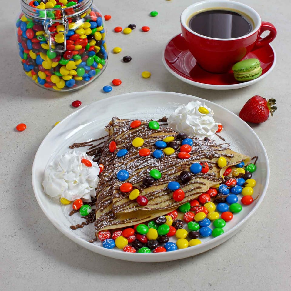 Chocolate crepe with M&M's, whipped cream, powdered sugar, a red coffee cup, and a green macaron.