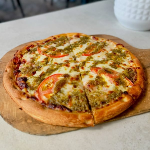 A freshly baked pesto pizza with mozzarella and sliced tomatoes on a wooden peel.
