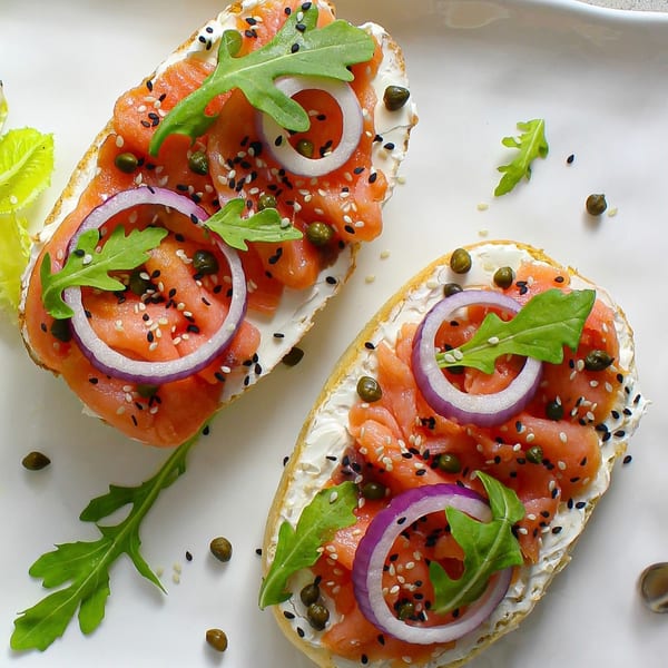 Two salmon and cream cheese bruschettas with red onion, arugula, capers, and sesame seeds.