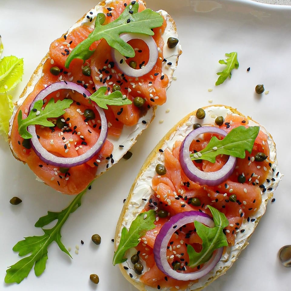 Two salmon and cream cheese bruschettas with red onion, arugula, capers, and sesame seeds.