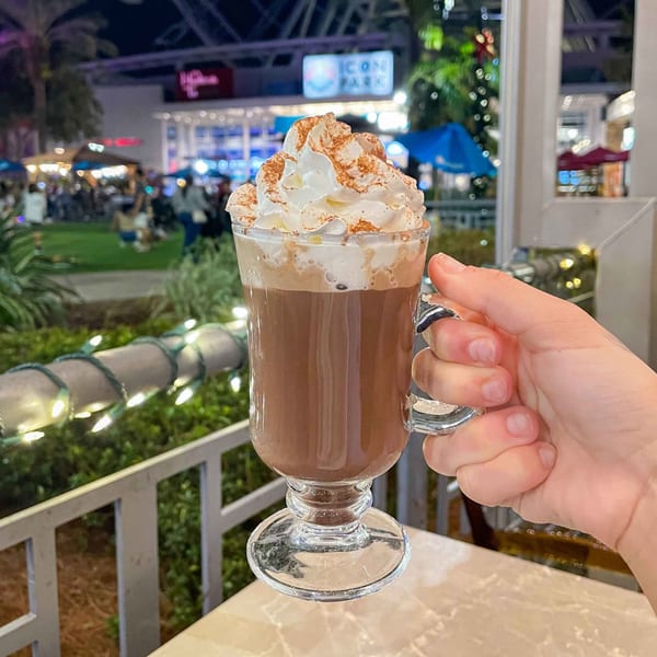 Hand holding a glass of hot chocolate with whipped cream and cinnamon at iCafe de Paris.
